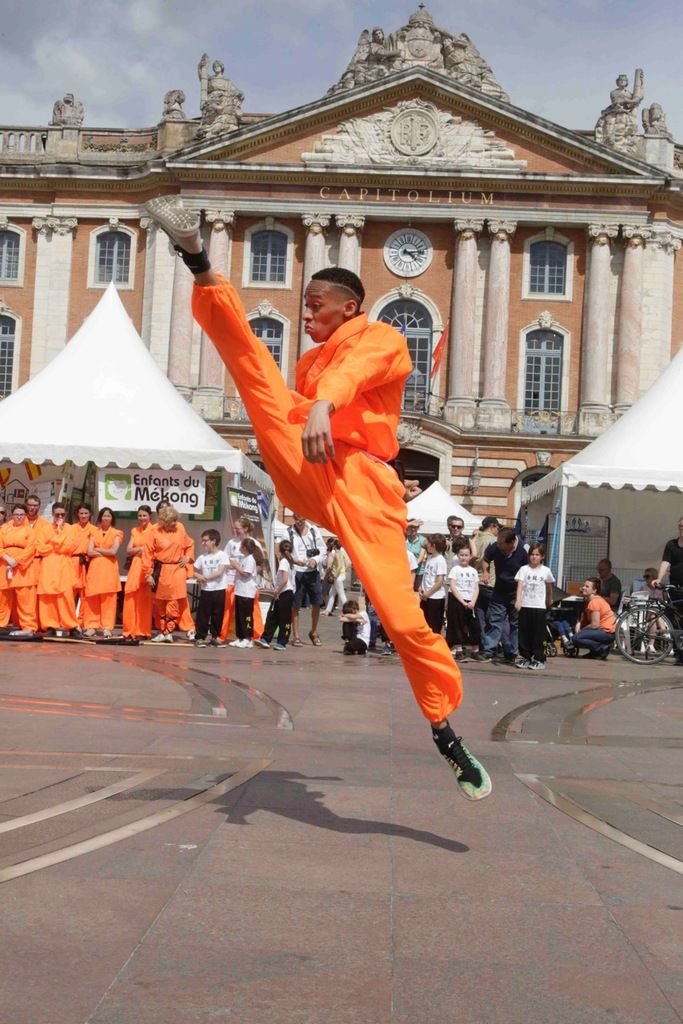 Un homme pratiquant le kung-fu sur la Place du Capitole à Toulouse, donnant un coup de pied haut, avec la façade emblématique du Capitole en arrière-plan.