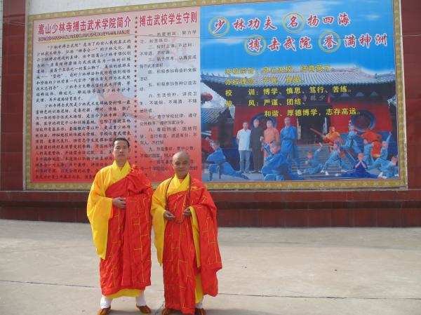 Deux moines Shaolin debout en méditation, vêtus de robes oranges traditionnelles, devant un bâtiment d'école.